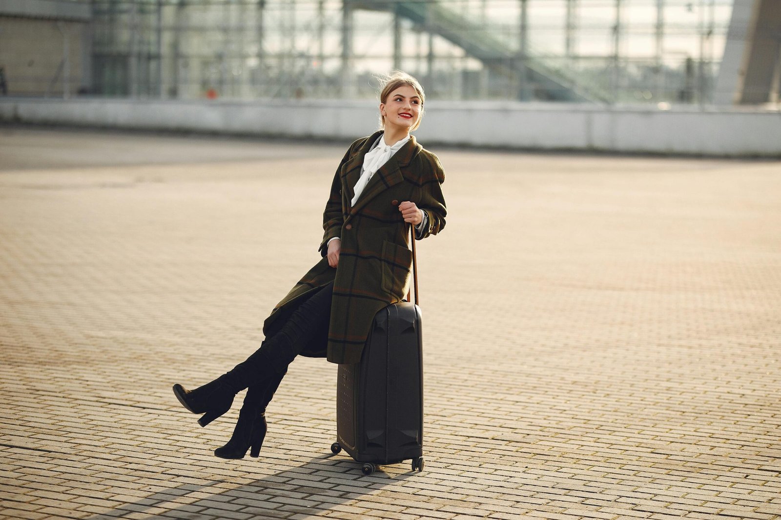 Cheerful stylish woman sitting on suitcase in front of contemporary glass building on urban street